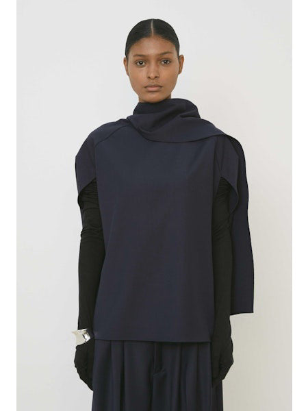 A woman with slicked-back hair and a neutral expression wears the MEIMEIJ Draped Cape Effect Top in navy over black long sleeves, paired with a matching navy skirt, set against a plain white background.