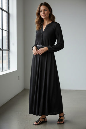 A woman wearing the MOMONI - AMEDE DRESS IN LUREX JERSEY and a black wide-brimmed hat stands in an ornately decorated room with large mirrors and intricate wall designs.