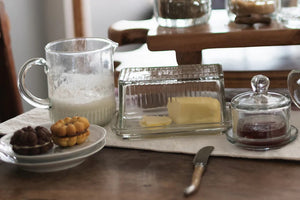 The ETUHOME - BISTRO GLASS MINI JUG by ETUHOME, filled with milk, rests on a white surface next to a butter dish and knife, with a clear glass baking dish and wooden container in the background.