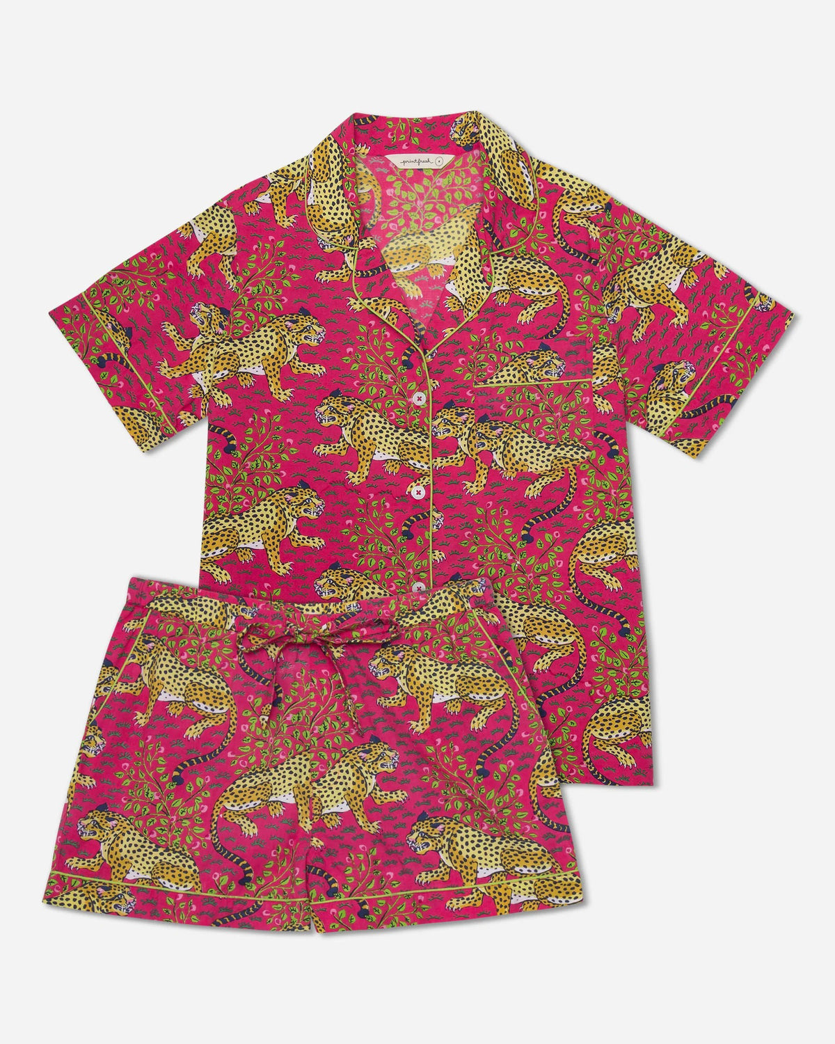 Against a beige backdrop, an individual smiles in their PRINTFRESH - BAGHEERA COTTON PAJAMA SHORT SET featuring a vibrant floral print, paired with fuzzy white slippers. Made from 100% organic cotton poplin, the long-wavy-haired person casually tucks their hands in their pockets.