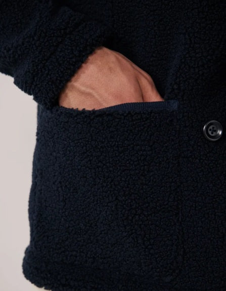 A young person with short dark hair wears the HARTFORD - Sherpa Utility Jacket in Navy by HARTFORD over a black shirt, looking directly at the camera against a plain light background. This warm sherpa layer brings both comfort and style to casual menswear.