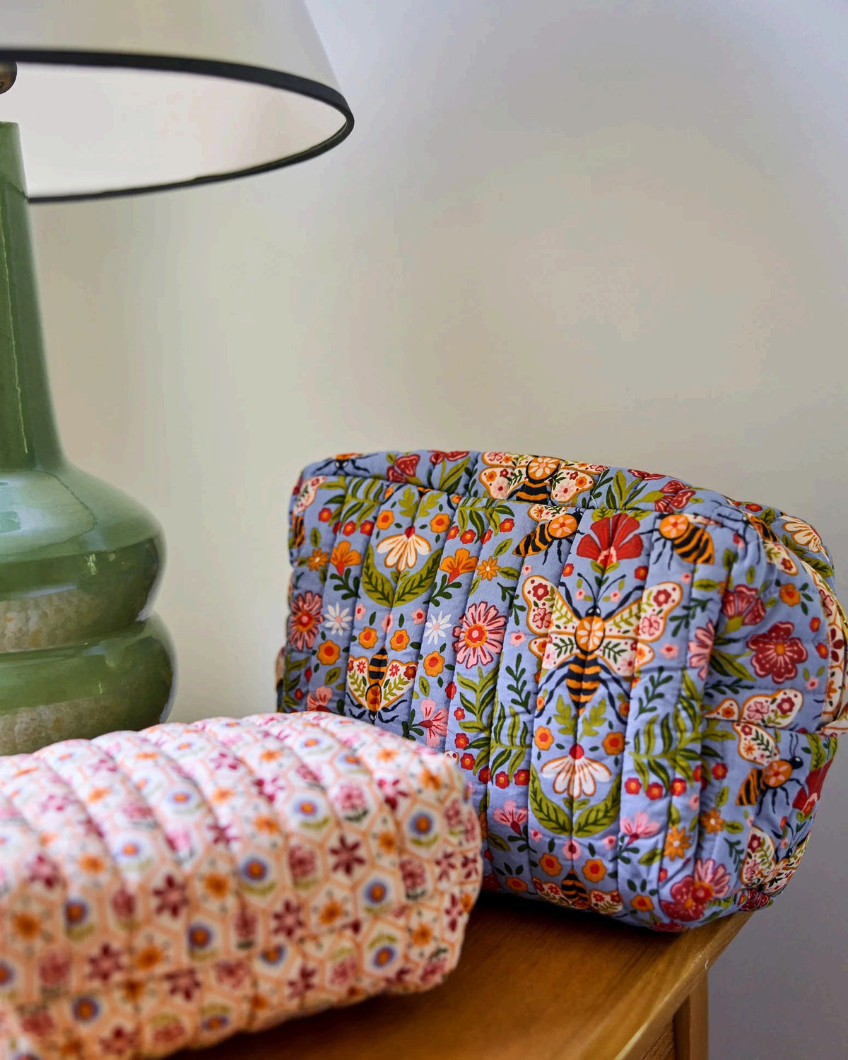 The PRINTFRESH Bee Tapestry Oversized Pouch Duo in Delphinium Blue features two quilted makeup pouches: the larger pouch showcases a blue bee and floral print, while the smaller pouch displays a cream background with red, yellow, and blue geometric shapes. Both PRINTFRESH bags are displayed on a tiled surface.
