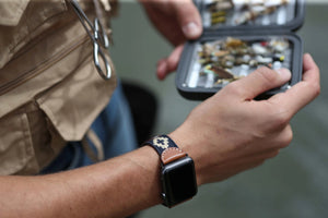The LA MATERA CORBINA Apple Watch Band in Space Black features a rectangular screen and artisan-crafted brown leather strap with a black woven band adorned with tan diamond patterns, showcasing elegance on a crisp white background.