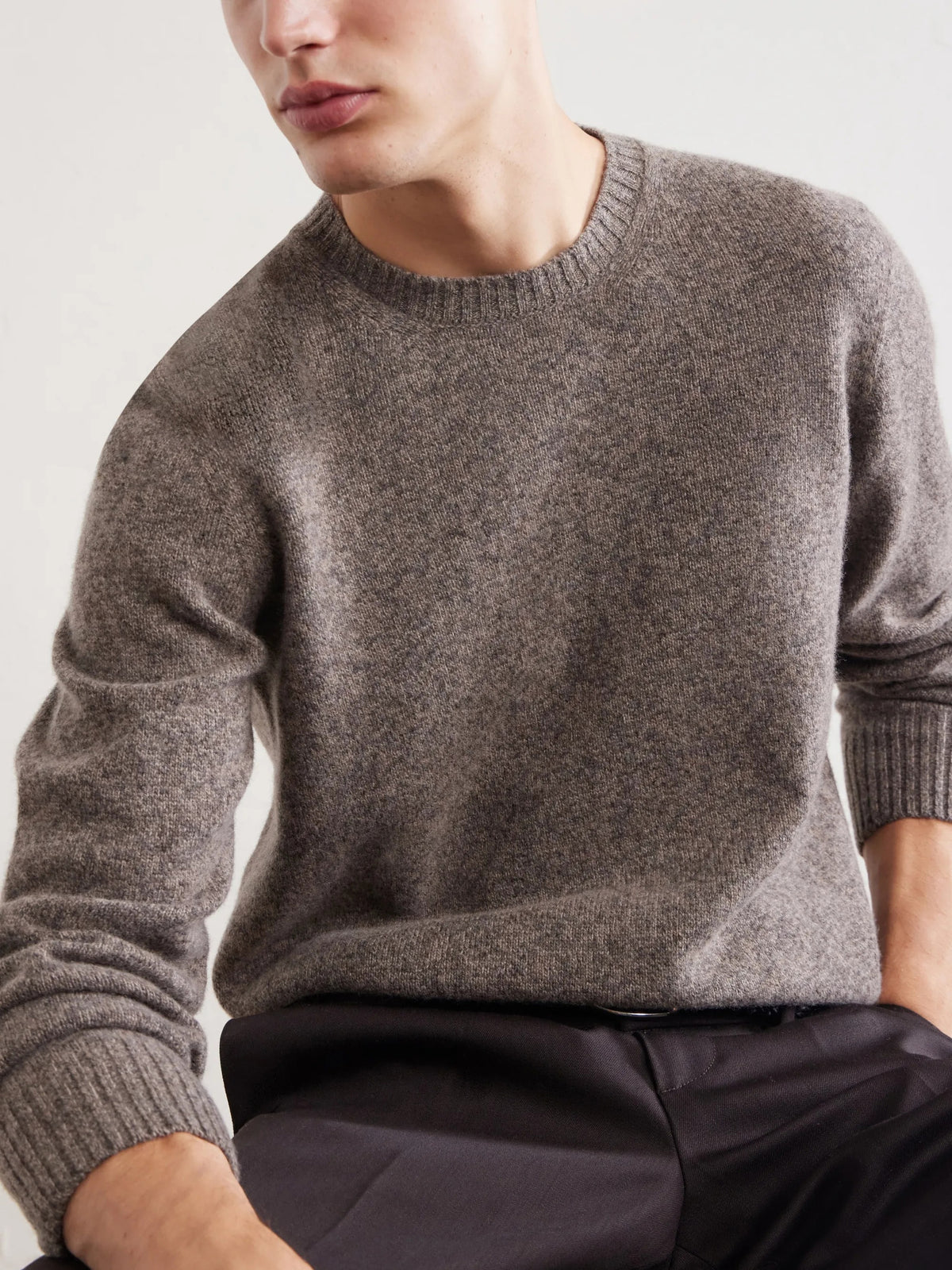 A person wearing the HARTFORD Seamless Wool Crew Pullover in Mushroom, an Italian-made sweater, paired with dark pants, is seated with one arm resting on their knee against a plain light background.