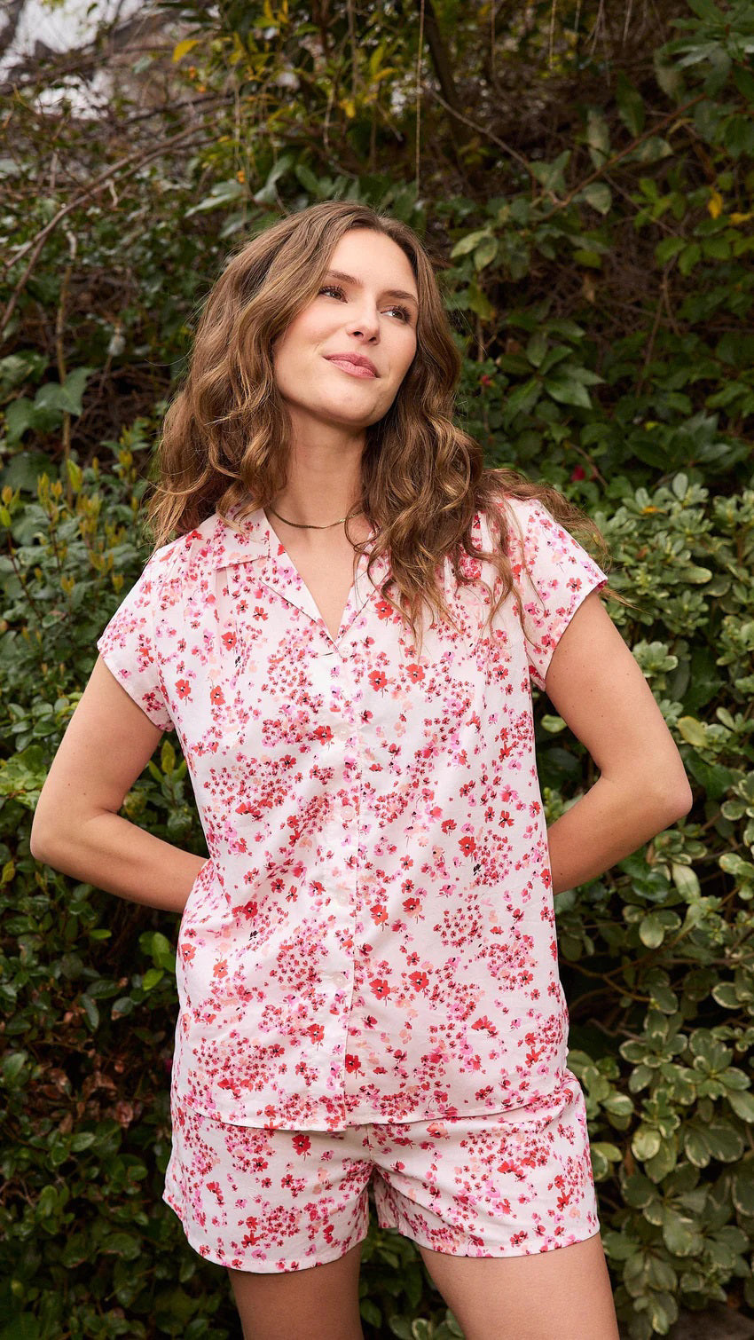 A smiling woman stands barefoot against a beige background, wearing the CAT&#39;S PAJAMAS - PETITE ROSETTE VOILE SHORT SET by CAT&#39;S PAJAMAS—a pink and white floral cotton voile pajama top and matching shorts—ideal for lightweight women&#39;s sleepwear.