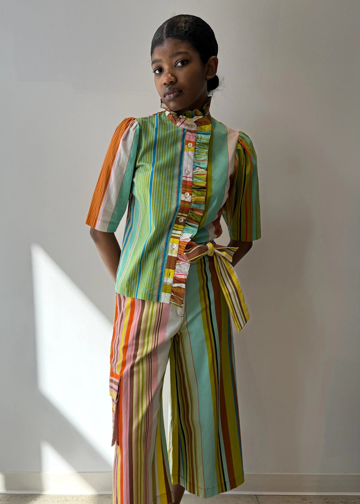 A person models the HOPE FOR FLOWERS Frilled Button Down Shirt in Combo Stripes by HOPE FOR FLOWERS, featuring vibrant vertical stripes and a ruffled collar, crafted from GOTS certified organic cotton for sustainability.