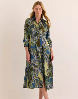 A woman stands against a plain light background, one hand in her pocket and smiling slightly, wearing the HINSON WU MARTINA 3/4 SLEEVE OPULENT PAISLEY PRINT DRESS—a collared midi dress crafted from fine Italian polyester in shades of blue, green, and olive.