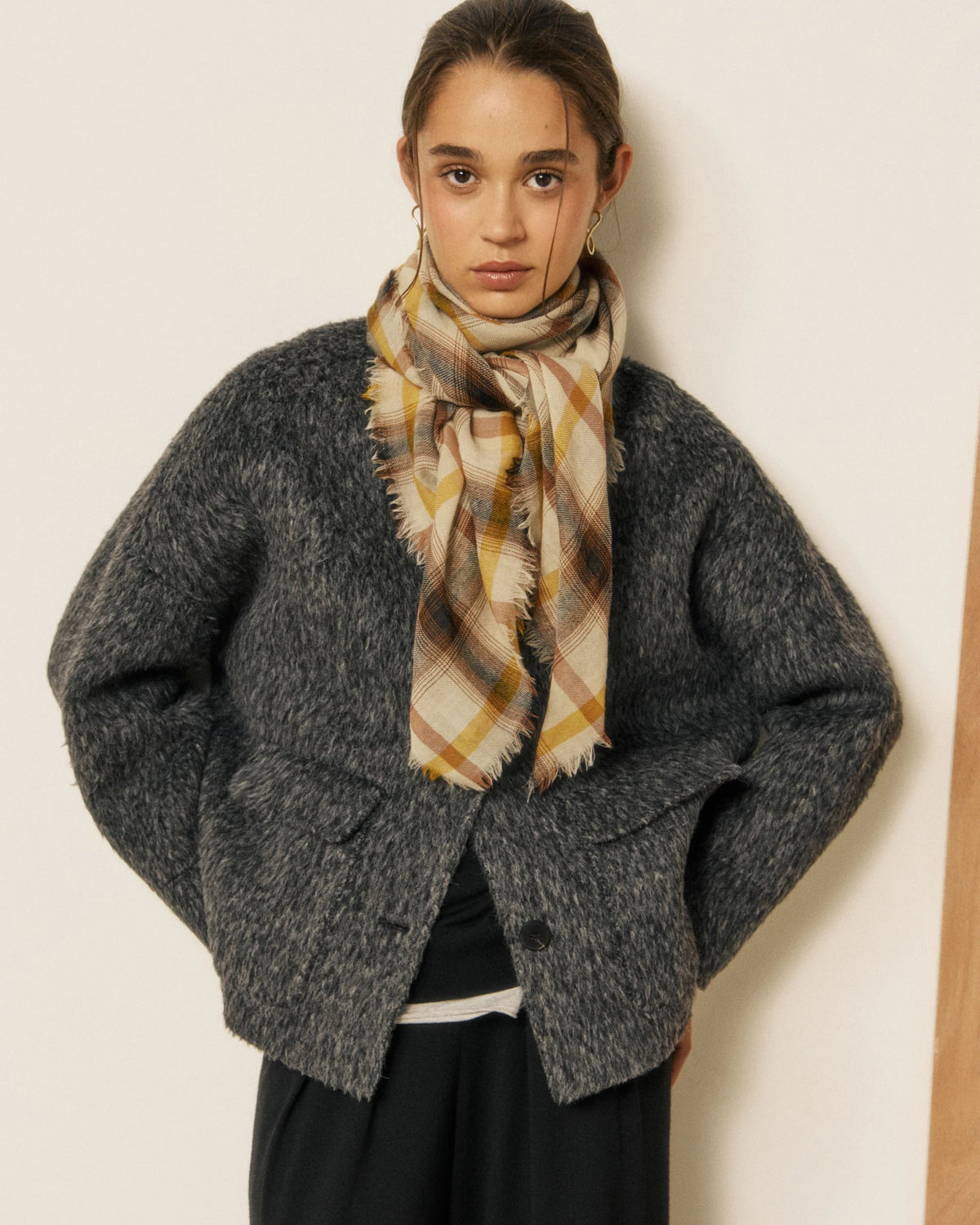 A young woman stands against a plain light background, looking directly at the camera with her hands in her pockets. She is wearing a fuzzy gray jacket, black pants, and the HARTFORD - CHECKED WOOL SCARF in yellow and brown by HARTFORD.