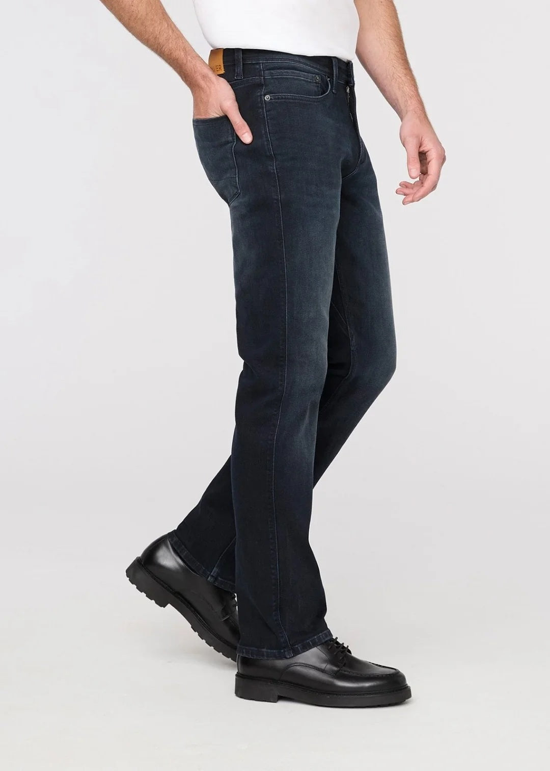 A man stands against a plain background wearing a black jacket, white t-shirt, DU/ER - PERFORMANCE DENIM+ SLIM STRIAGHT PANTS IN CARBON STONE, and black shoes, with one hand in his pocket and a neutral expression.