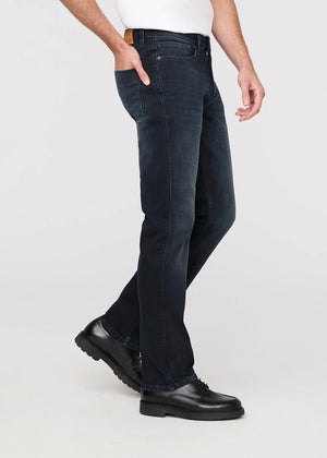 A man stands against a plain background wearing a black jacket, white t-shirt, DU/ER - PERFORMANCE DENIM+ SLIM STRIAGHT PANTS IN CARBON STONE, and black shoes, with one hand in his pocket and a neutral expression.