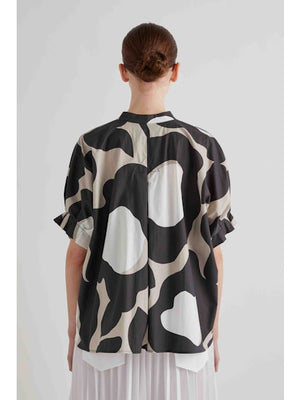 A person in dark sunglasses and a loose-fitting MEIMEIJ FRANK AND EILEEN MOONBEAM TOP with a bold black and beige abstract pattern, paired with a white pleated skirt, stands against a plain light background.