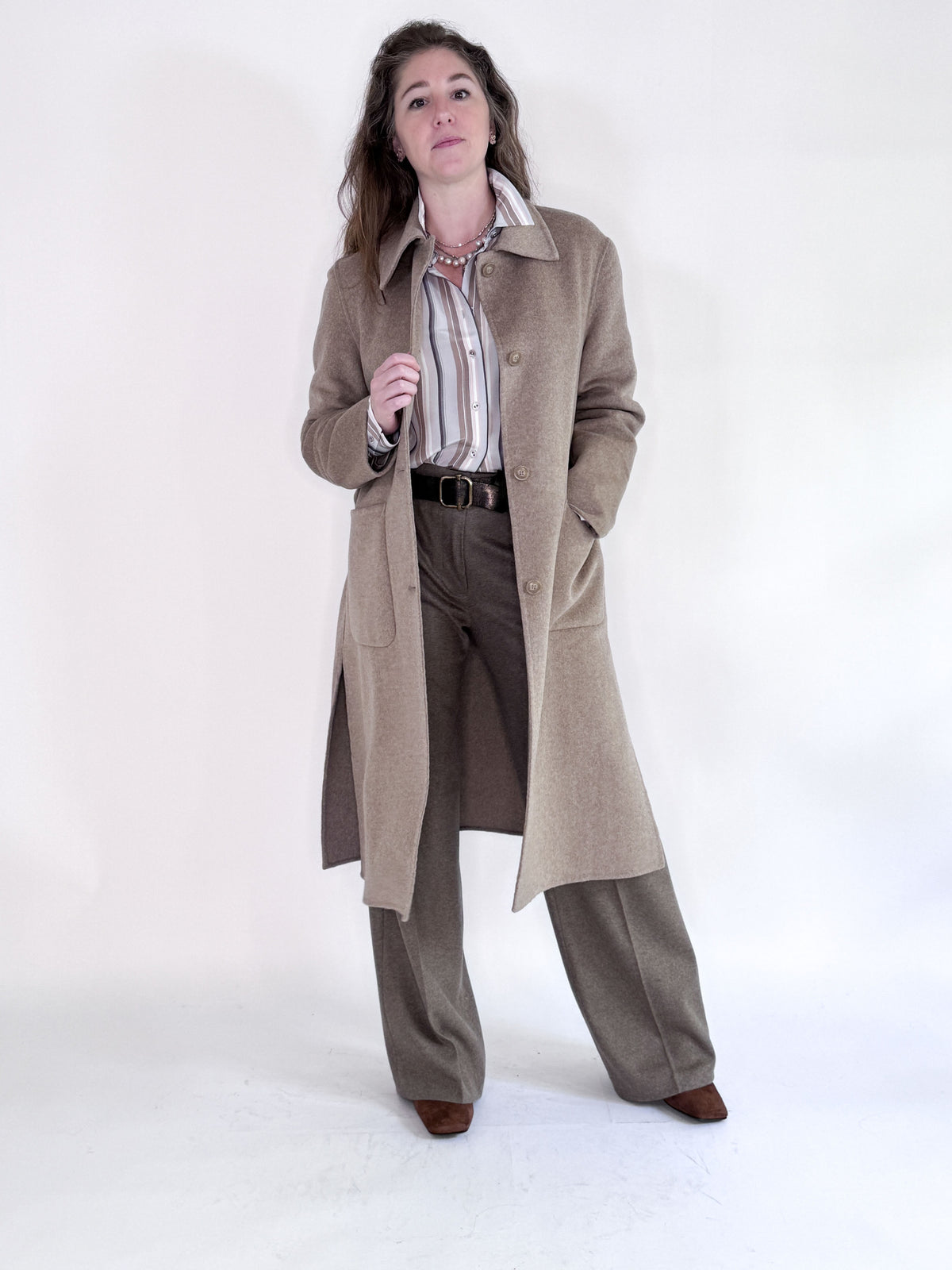 A woman poses confidently with one hand in her pocket, wearing the SEVENTY VENEZIA - WOOL BLEND LONG COAT by SEVENTY over a striped shirt, brown trousers, and brown shoes against a plain background.
