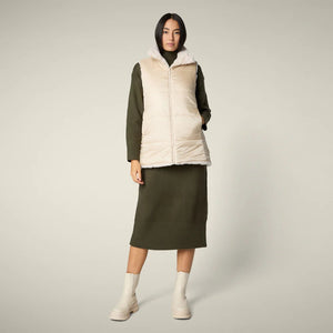 A woman with straight blonde hair wears the SAVE THE DUCK NIVES REVERSIBLE VEST over a white long-sleeve top, beige wide-leg pants, and black shoes, standing against a plain background and highlighting the vest's chic style.
