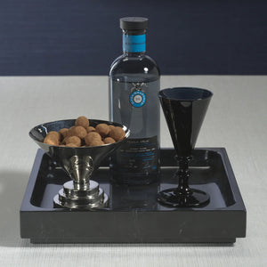 A black bottle of tequila sits on a ZODAX NERO MARQUINA NATURAL MARBLE TRAY - LARGE, accompanied by a black goblet and bowl of chocolate treats, all arranged on a light surface with a dark backdrop.
