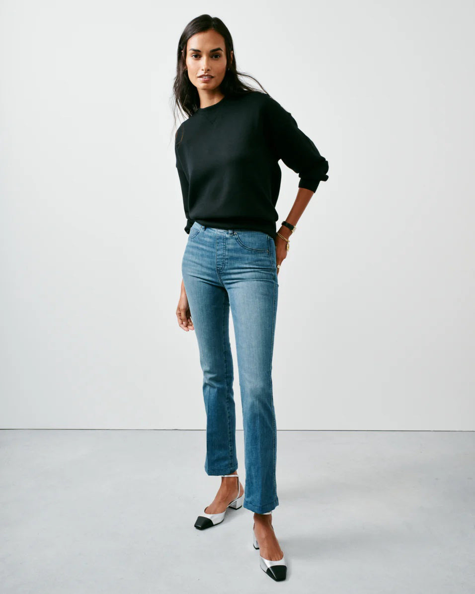 A person stands against a plain backdrop, confidently showcasing their style in SPANX's KICK FLARE JEAN, made with 4-way stretch denim. They pair it with a black sweater and black-and-white heels, their long dark hair cascading as they pose with one hand in their pocket.