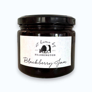 The FEARRINGTON SIGNATURE COLLECTION - FEARRINGTON HOUSEMADE BLACKBERRY JAM features a stylish black and white striped ribbon tied around the lid. The label reveals ingredients including blackberry, vanilla, lemon, pectin, and sugar. This handcrafted jam is made with meticulous care and is an ideal hostess gift. Please be aware that it may contain allergens.