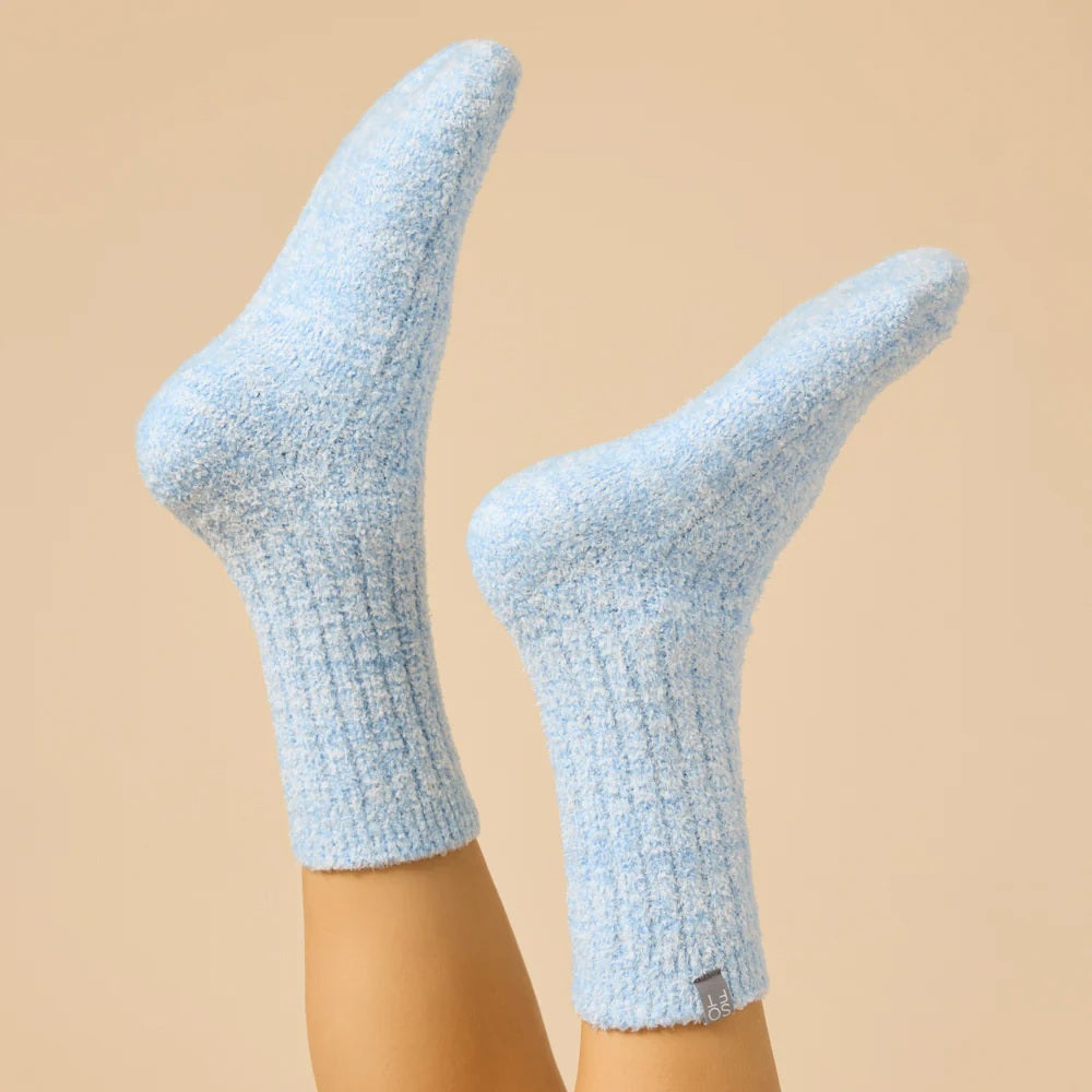 Two feet wear SOFTIES - MARSHMALLOW CREW SOCKS IN HEATHER SKY BLUE, featuring a textured pattern and small gray tag near the cuff, raised against a beige background.