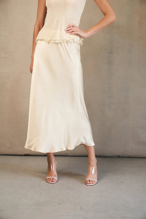 A woman models the ROSE & CROWN - SATIN MIDI SKIRT in silky blue, paired with a short-sleeved light blue knit top, standing against a white background. Her head is not visible in the image.