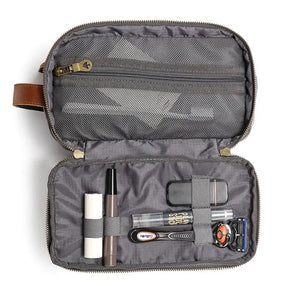 The TSD BRAND - Turtle Ridge Toiletry Bag in grey features waxed canvas, leather details, and a zip closure. It is open to show neatly organized travel essentials inside—bottles, a tube, a brush, and a white rectangular item.