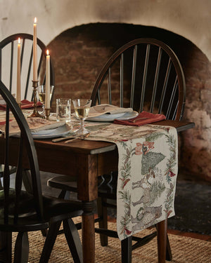 A CHRISTMAS CHEER EMBROIDERED LINEN TABLE RUNNER featuring woodland animals in winter attire and Christmas trees, draped over a wooden dining table set with plates, glassware, and candlesticks.