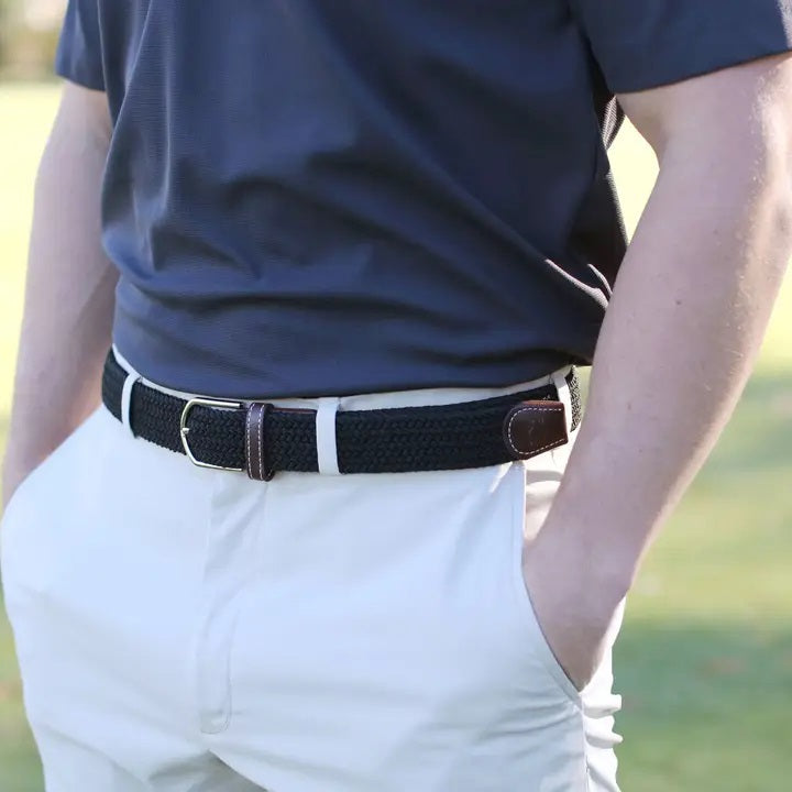 Outdoors, a person with hands in pockets wears a navy blue shirt, light-colored pants, and the ROOSTAS - BETHPAGE WOVEN STRETCH BELT by ROOSTAS featuring brown leather accents.