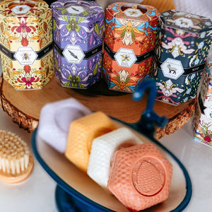 A decorative BAUDELAIRE - GOATS MILK HONEY SOAP 1.4 OZ QUARTET box sits beside a stack of four pale pink, hexagon-shaped soap bars. The floral-patterned box features an illustration of a goat, drawing attention to this gentle triple-milled soap from BAUDELAIRE, crafted with nourishing goat milk and natural honey.