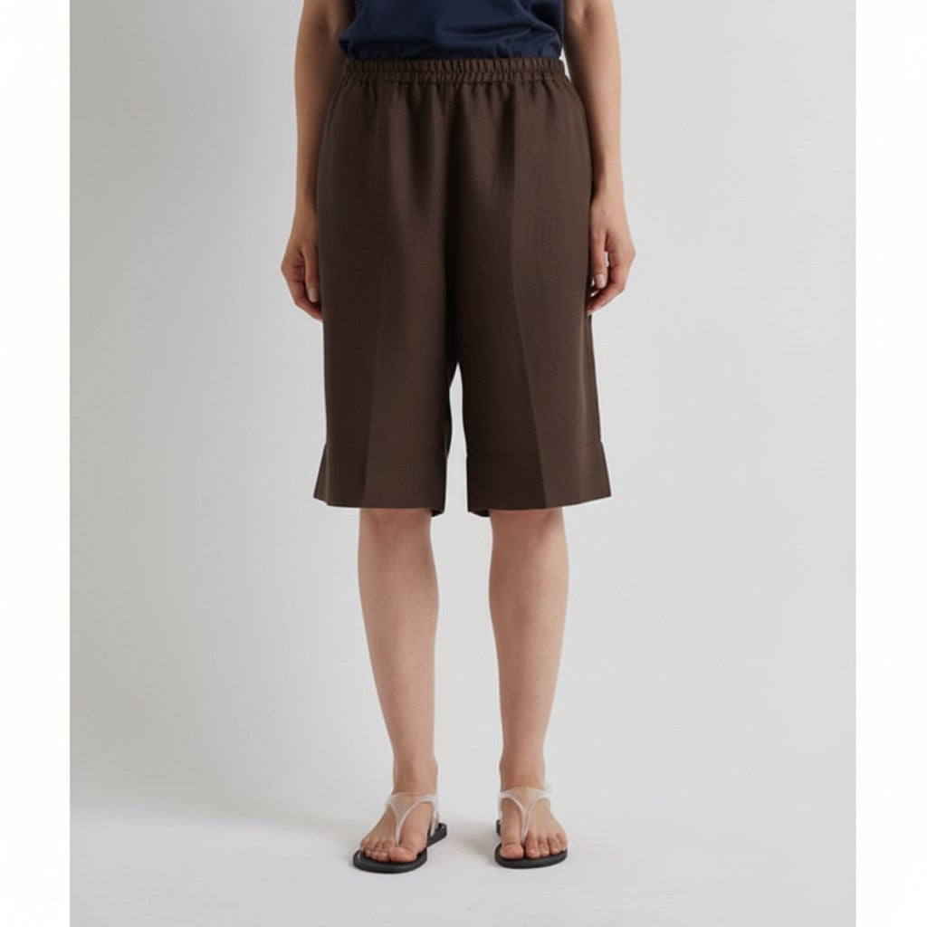 A person wears MEIMEIJ's Linen Shorts in Mocha and clear strap sandals with a navy top, standing against a light background, embodying relaxed summer tailoring.