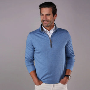 A man wearing the COLLARS AND CO - HEATHER CLOUD COTTON 1/4 ZIP SWEATER IN MEDIUM BLUE WITH GREY TRIM over a white collared shirt stands with his hands in his pockets, smiling and looking to the side against a plain gray background.
