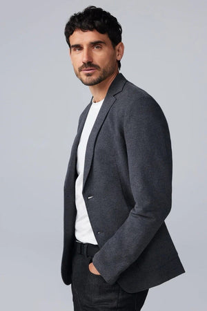 A man fastens the MACK WELDON Ace Blazer in Charcoal Heather over a gray hoodie, white tee, and dark jeans, paired with white sneakers with beige soles. He stands against a plain background, looking slightly to the side.