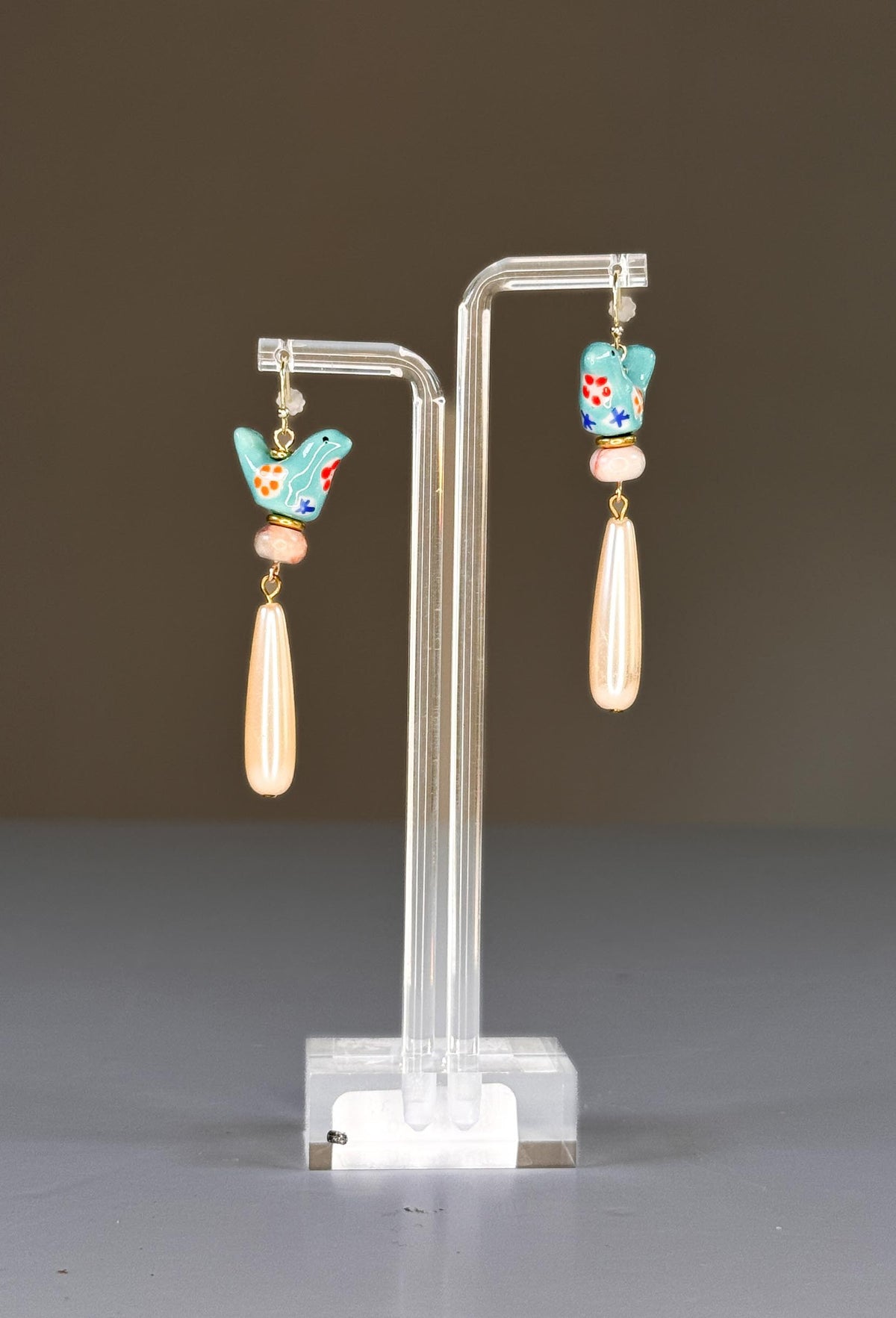 The SANDRINE FRANCE STUDIO - GEMMA EARRINGS feature turquoise ceramic bird charms with painted flowers, beige round beads, and long teardrop-shaped pearl dangles on gold hooks, shown against a white background.