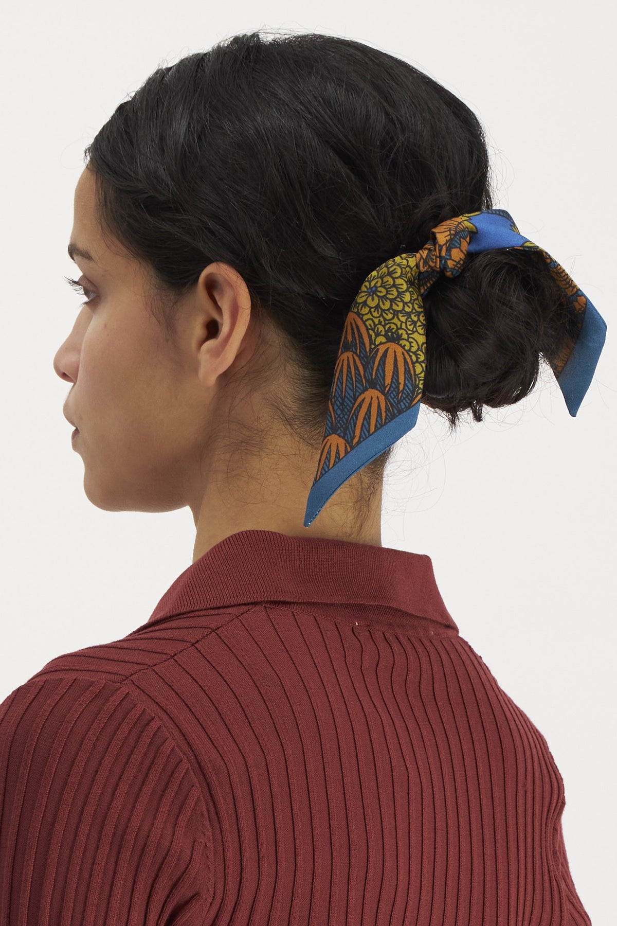 A person with dark hair styled in a low bun, adorned with the INOUI EDITIONS - MAGNUS SMALL SILK RIBBON featuring orange and green designs on a blue base, wearing a ribbed, long-sleeved maroon top. The timeless accessory from INOUI, made from 100% silk, is shown from the side against a plain white background.