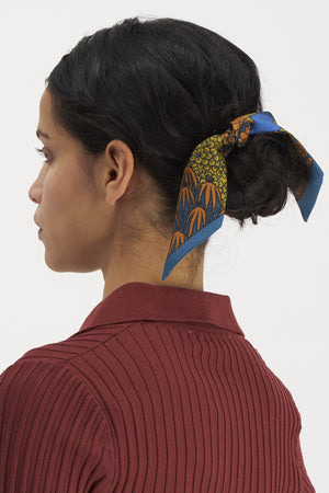 A person with dark hair styled in a low bun, adorned with the INOUI EDITIONS - MAGNUS SMALL SILK RIBBON featuring orange and green designs on a blue base, wearing a ribbed, long-sleeved maroon top. The timeless accessory from INOUI, made from 100% silk, is shown from the side against a plain white background.