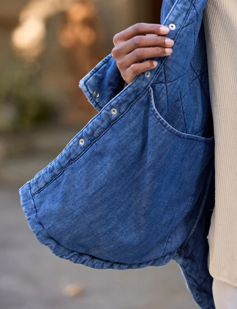 A person holds open a blue FRANK &amp; EILEEN - EILEEN Quilted Shirt Jacket in Famous Denim Distressed Indigo Wash, revealing its interior pocket and distinctive diamond quilted pattern. The person is wearing a cream-colored, ribbed top, with a blurred background.