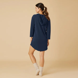 A woman stands on a beige background wearing the SOFTIES Terry Hooded Lounger in Midnight, paired with light gray socks. With her hair tied back and a slight smile, she showcases the brand's signature resort-ready comfort in this stylish look.