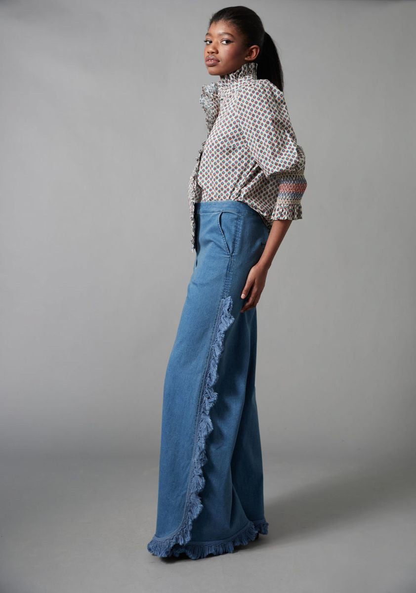 A woman models the HOPE FOR FLOWERS - FRILLED BUTTON DOWN SHIRT by HOPE FOR FLOWERS, made from GOTS certified organic cotton, with a high collar and wide sleeves, paired with flared blue jeans featuring fringe along the side seams.