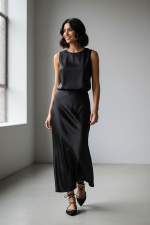A woman stands smiling, wearing the MILVA MI - LONG SATIN SKIRT IN BLACK by MILVA, styled with a fluid silhouette. The skirt flows to the floor in lustrous black satin, paired with a fitted top that features long sleeves and a cowl neckline. She has wavy brown hair and wears black shoes, standing against a plain light background.