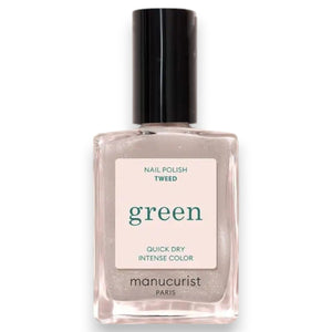 A hand painted with MANUCURIST - GREEN™ NAIL POLISH IN TWEED by MANUCURIST INC, a short pearly grey shade, rests on a shiny red surface. The person wears a gold ring featuring a large green stone on their middle finger, while a green and white bowl is partially visible in the corner.