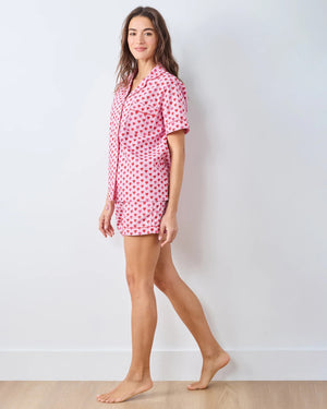 A person wearing the PRINTFRESH - MINI CANDY HEARTS PAJAMA SHORT SET by PRINTFRESH, featuring pink fabric with red heart patterns, stands with one hand in their shorts pocket. The image highlights the details of the pajama set and does not show the person's face.