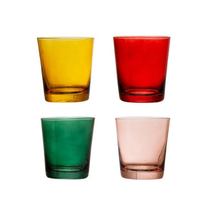 Three colorful CREATIVE COOP LOW BALL GLASSWARE pieces (red, yellow, green) are stacked next to a gold cocktail shaker with a red top, decorative ornaments, and a bar spoon, with a small tree in the background.