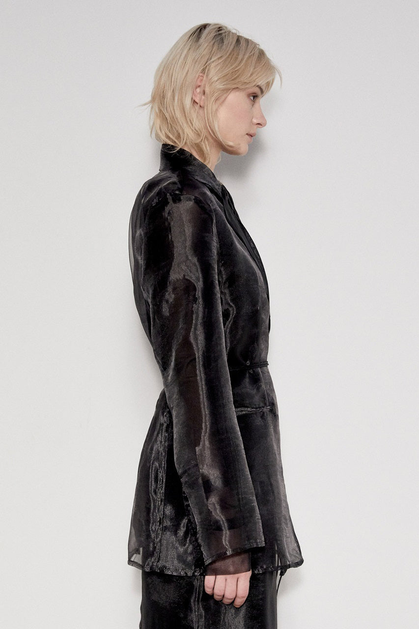 A person with blonde hair stands against a plain background, wearing the MEIMEIJ - SHEER JACKET by MEIMEIJ, which features long sleeves, a subtle sheen, and a tie at the waist. They have a neutral expression, embodying contemporary minimalist style. To elevate the look subtly, pair it with denim and a t-shirt underneath.