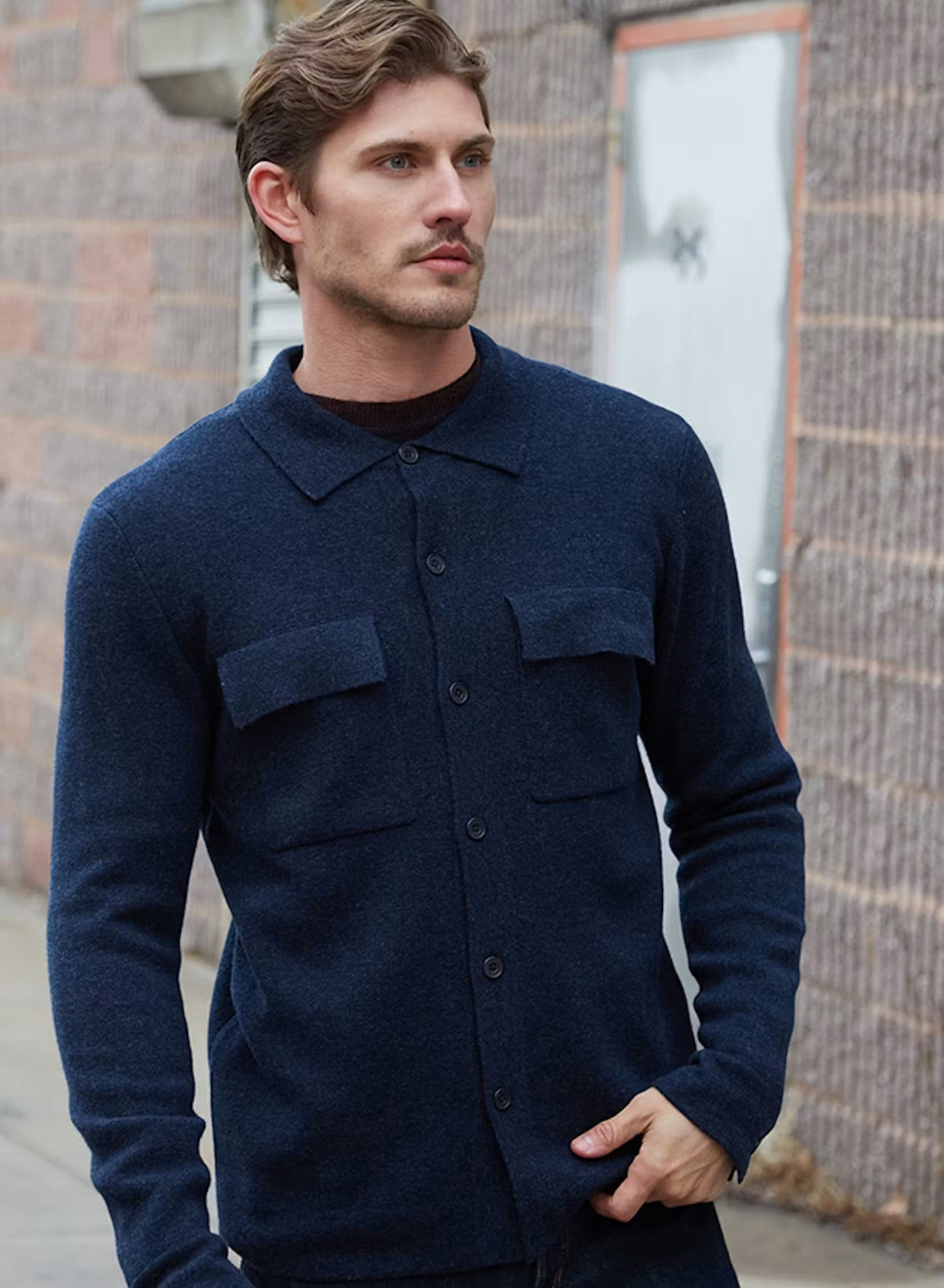 A man wearing the AUTUMN CASHMERE Milano Stitch Shacket in Midnight, featuring two chest pockets, stands with one hand in his pocket and looks to the side against a white background.
