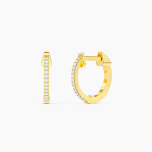 A woman with blonde hair styled in a low bun wears the ELLA STEIN - HALF HEARTED ADVENTURE NEVER HOOP EARRINGS from ELLA STEIN, featuring small gold plated hoops adorned with sparkling stones. The photo captures her profile against a neutral background.