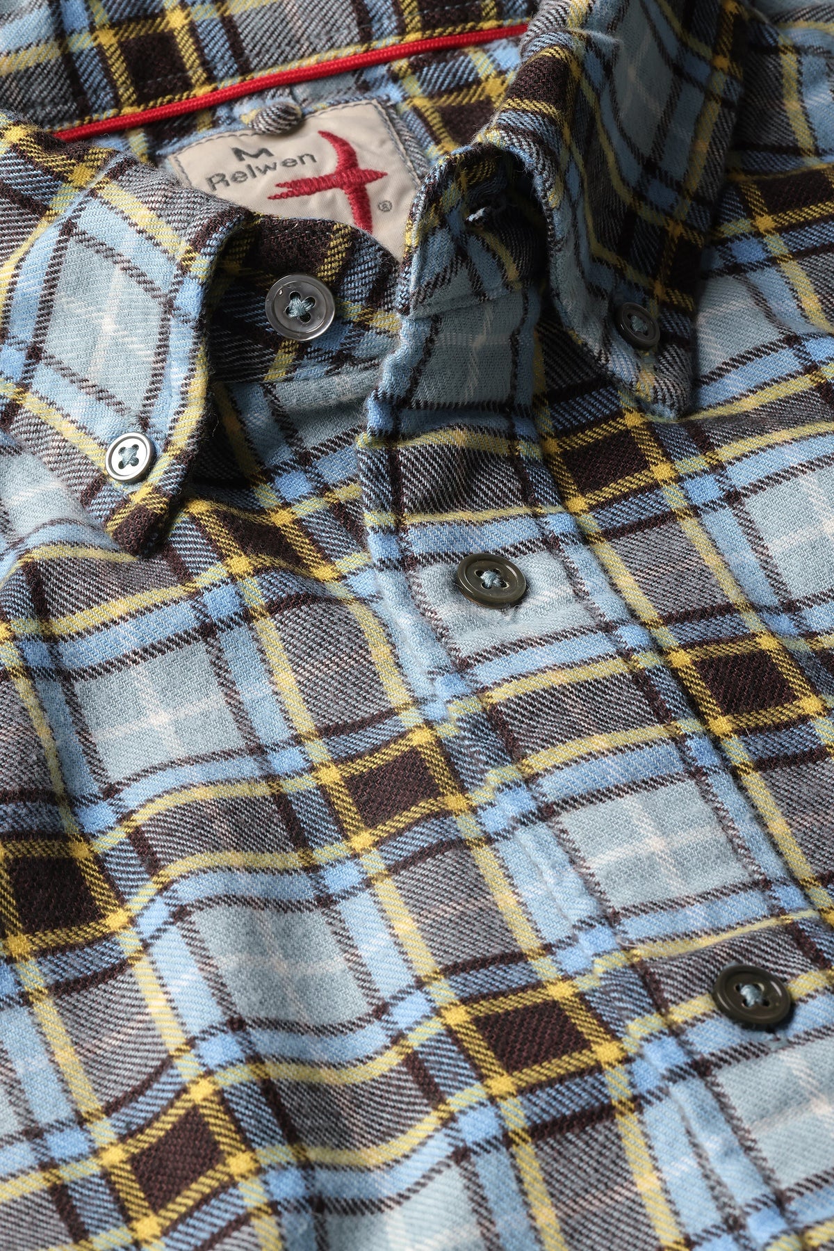 A close-up of the RELWEN Flyweight Flannel Shirt in Pale Blue/Brown Multi shows a blue, brown, and yellow plaid pattern with gray buttons. Made from lightweight, yarn-dyed cotton, this shirt features a partially visible RELWEN label at the top.