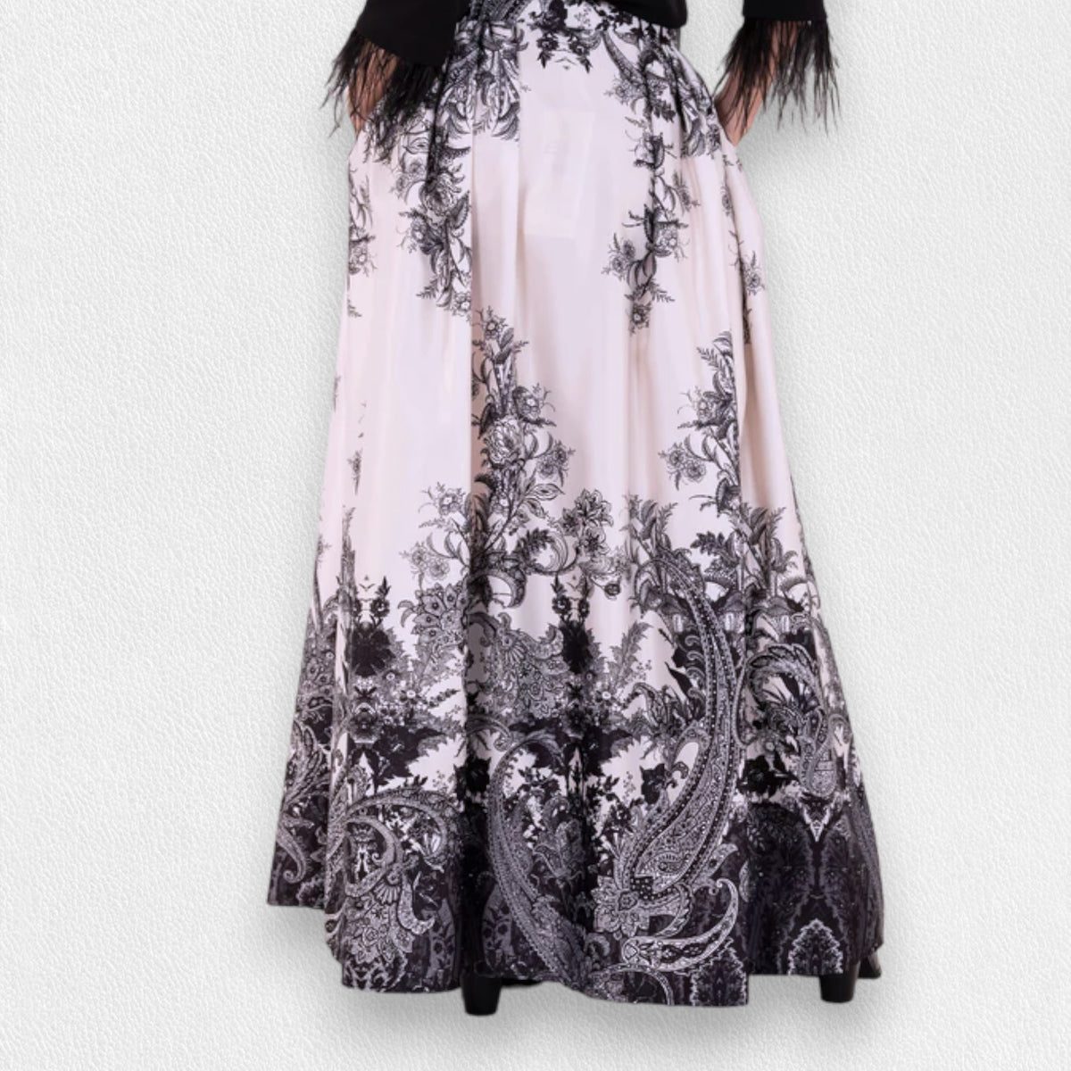 A woman stands against a plain gray backdrop wearing a sheer black blouse with ruffle details at the chest and MILVA&#39;s LONG MAXI SKIRT, featuring a detailed black-and-white pattern. The high-waisted, floor-length skirt, with welt pockets and a side closure with concealed zip, cinches at her gathered waist. She wears black shoes.