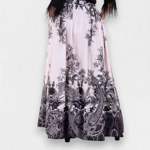 A woman stands against a plain gray backdrop wearing a sheer black blouse with ruffle details at the chest and MILVA's LONG MAXI SKIRT, featuring a detailed black-and-white pattern. The high-waisted, floor-length skirt, with welt pockets and a side closure with concealed zip, cinches at her gathered waist. She wears black shoes.