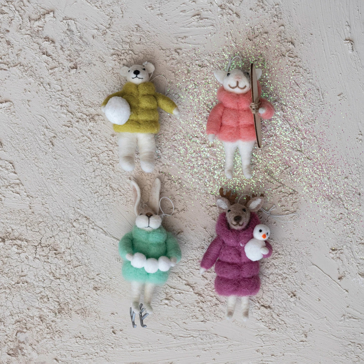 The CREATIVE COOP HANDMADE WOOL FELT ANIMAL IN WINTER PUFFER COAT ORNAMENT set features four animals—rabbit with skates, polar bear with snowboard, reindeer with penguin, and polar bear with snowball—each hanging by a string.