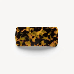 A person with brown wavy hair in a low twist, secured by the MACHETE - JUMBO BOX BARRETTE IN TORTOISE, is seen from behind wearing gold hoop earrings and a black top.