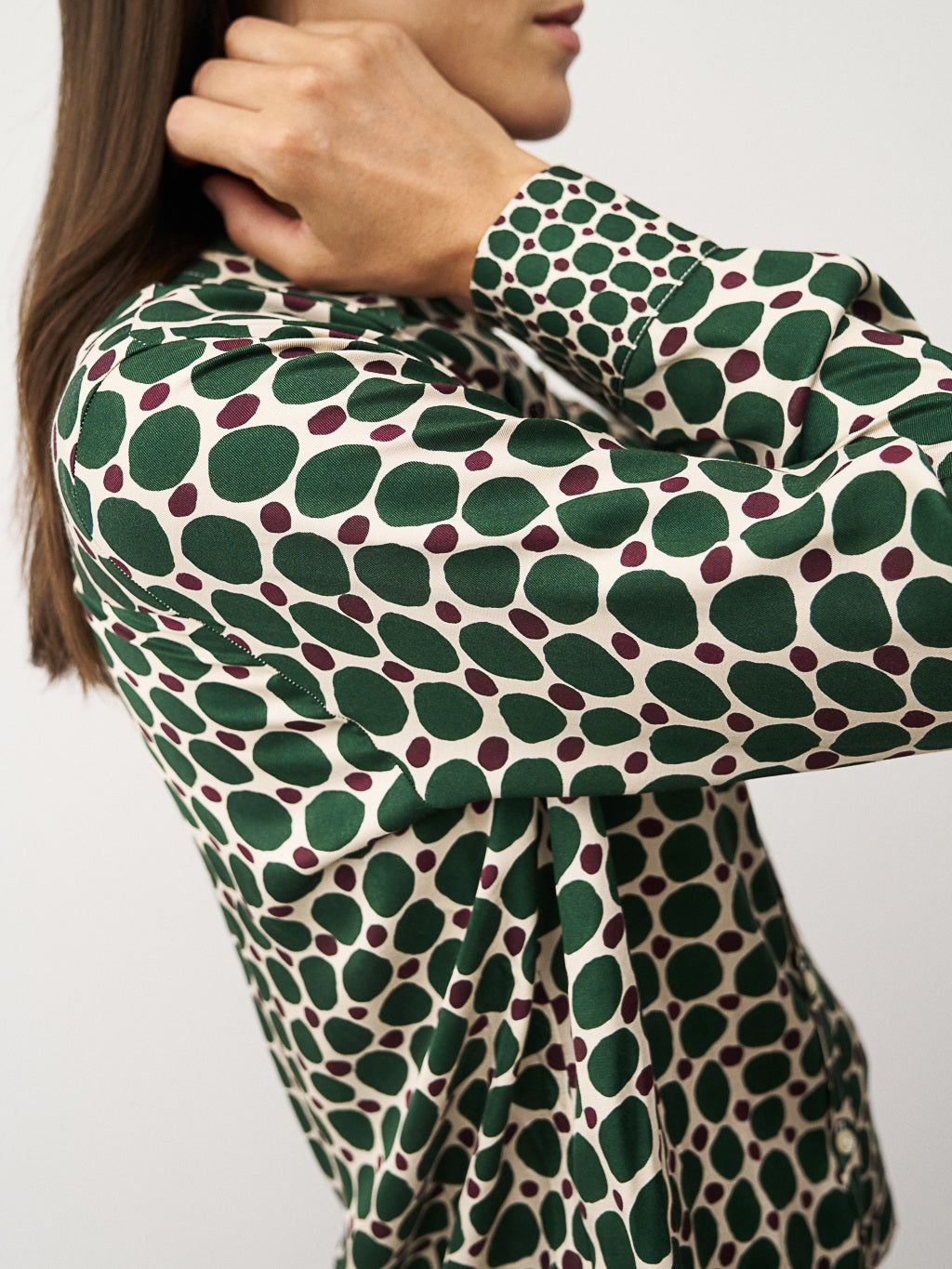 A woman with long brown hair smiles gently, arms crossed, wearing the 0039 ITALY Sia Blouse—a green and white geometric print blouse with pink dots—paired with brown pants against a plain white background.