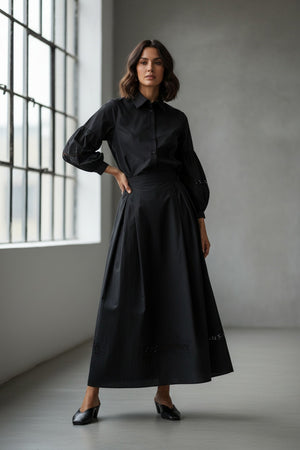 The MILVA MI Cotton Midi Skirt in Black by MILVA features a solid, subtly textured surface with slight shade variations and an elegant embroidered cutwork hem.
