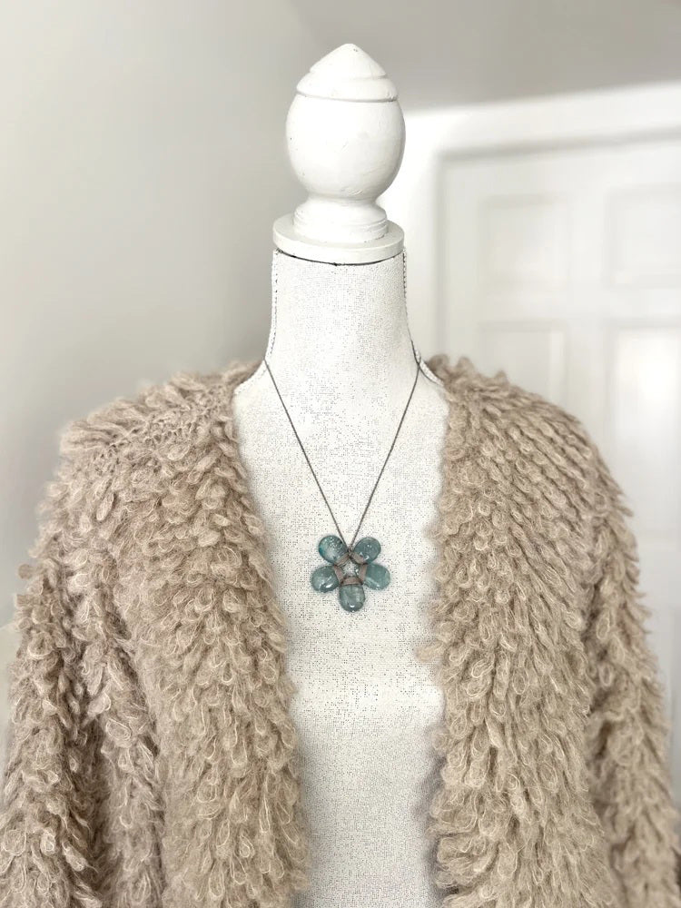A mannequin in a textured beige cardigan showcases the ANN LIGHTFOOT - BLUE CALCITE FLOWER NECKLACE, highlighted by a striking blue-green pendant. A softly lit white door appears in the background.